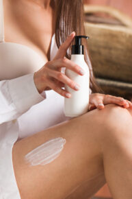 A close-up of a moisturizer on a woman's leg, in the other hand she holds a mockup of a white dispenser with cosmetics. Body care, skin tone.